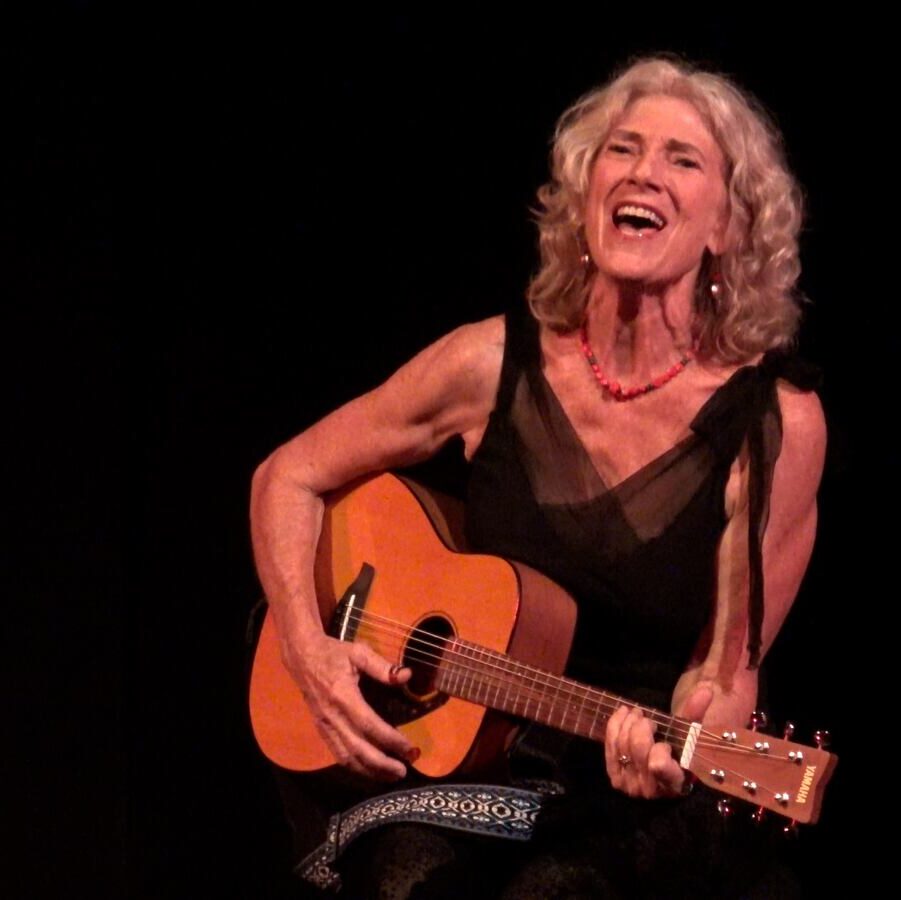 Silver-haired woman playing guitar on stage with a dark background